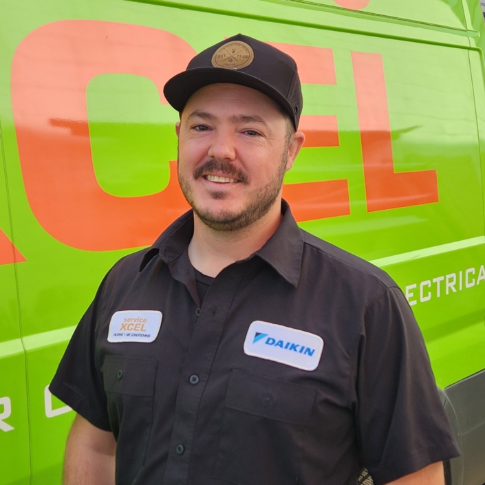 Smiling ServiceXEL technician in branded uniform and hat standing in front of green and orange company van